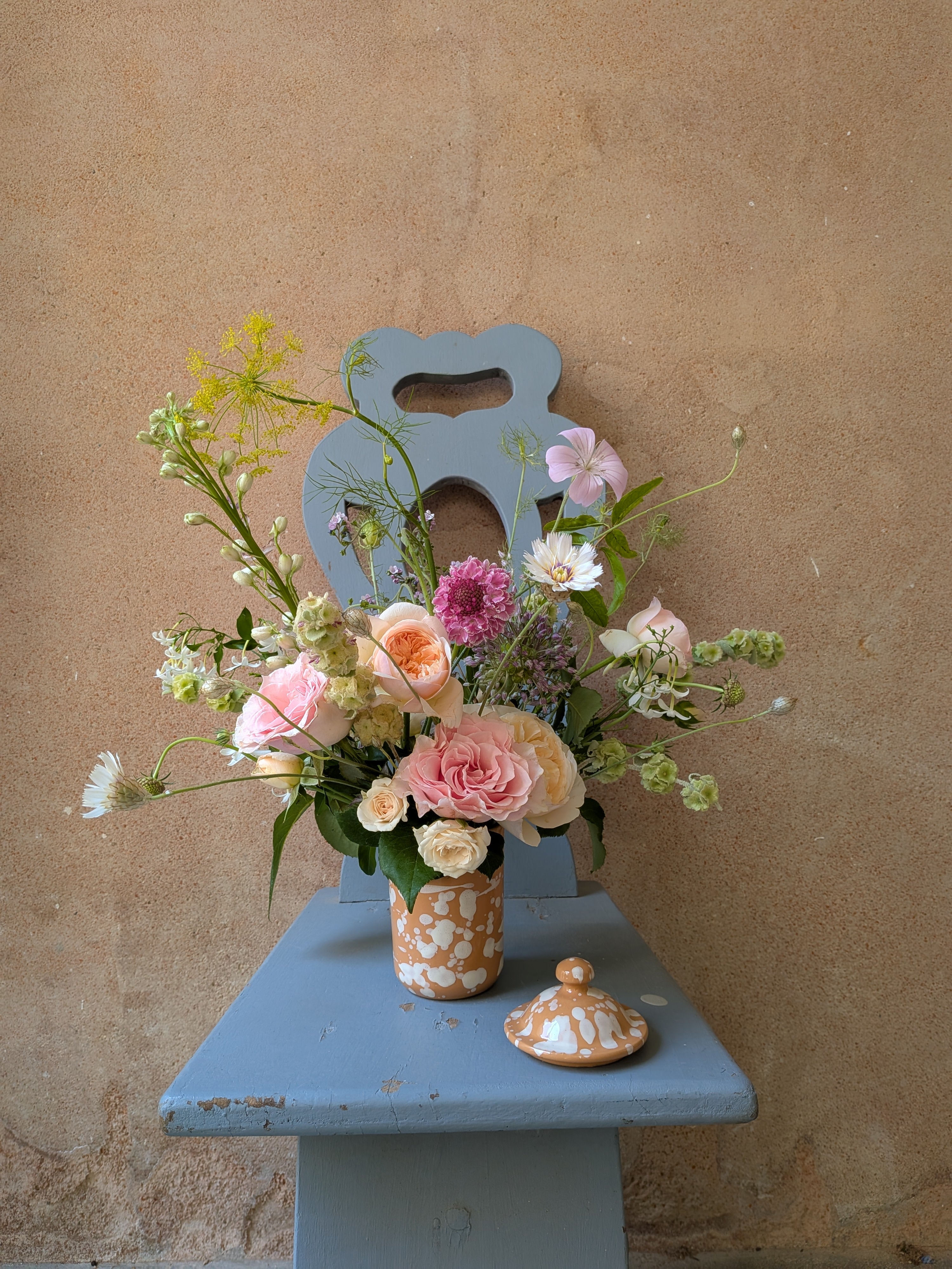 Floral arrangement in a decorative vase on a small blue chair against a beige wall.